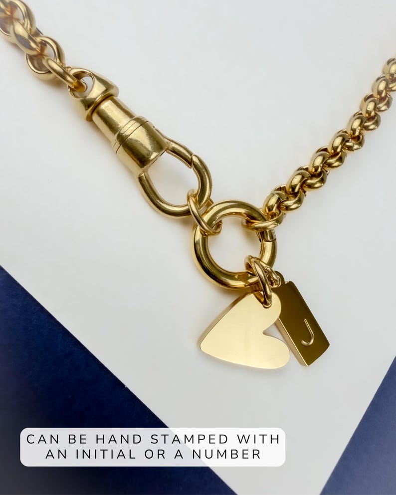 May include: A gold-toned chain necklace featuring a swivel clasp, a circular ring, and two charms: one heart-shaped and one rectangular with the letter J. The text CAN BE HAND STAMPED WITH AN INITIAL OR A NUMBER is visible at the bottom.