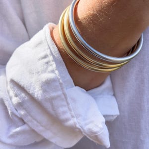May include: A collection of gold and silver bangles stacked on a wrist, set against a white linen shirt. The bangles vary in color, with some appearing gold and others silver. The shirt's sleeve is partially visible, with the cuff turned up.