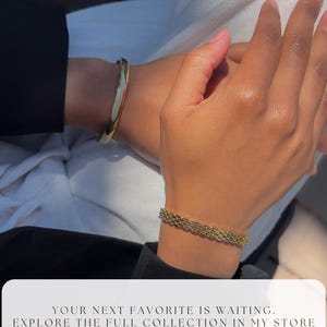 May include: A close-up of two wrists adorned with gold bracelets. One is a solid, polished bangle, and the other is a chain-link style. The image includes the text: "YOUR NEXT FAVORITE IS WAITING. EXPLORE THE FULL COLLECTION IN MY STORE FOLLOW LINK IN DESCRIPTION."
