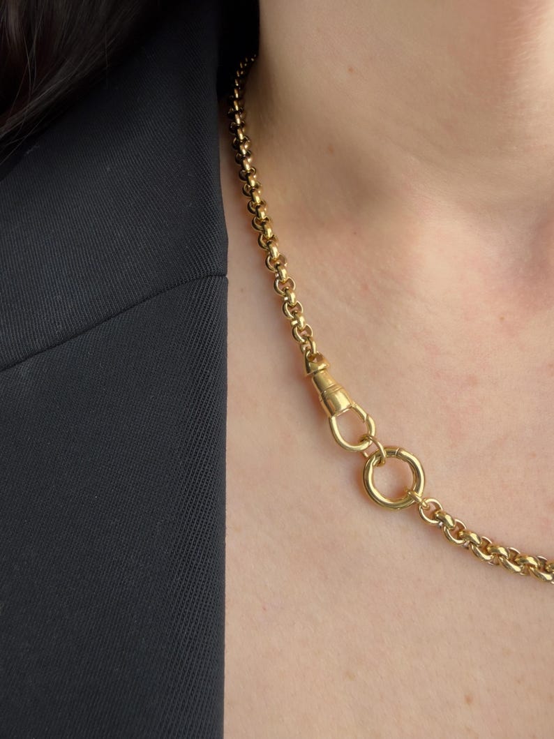 May include: A gold-toned chain necklace featuring round, interlocking links. The necklace includes a prominent swivel clasp mechanism connected to a circular ring, allowing for the attachment of pendants or charms along the chain length.