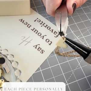 May include: A gold-colored heart charm being attached to a gold chain with metal pliers. A cream-colored card with the text 'say Hi to your new favorite!' is visible, along with a small dish containing various gold-colored jewelry components.