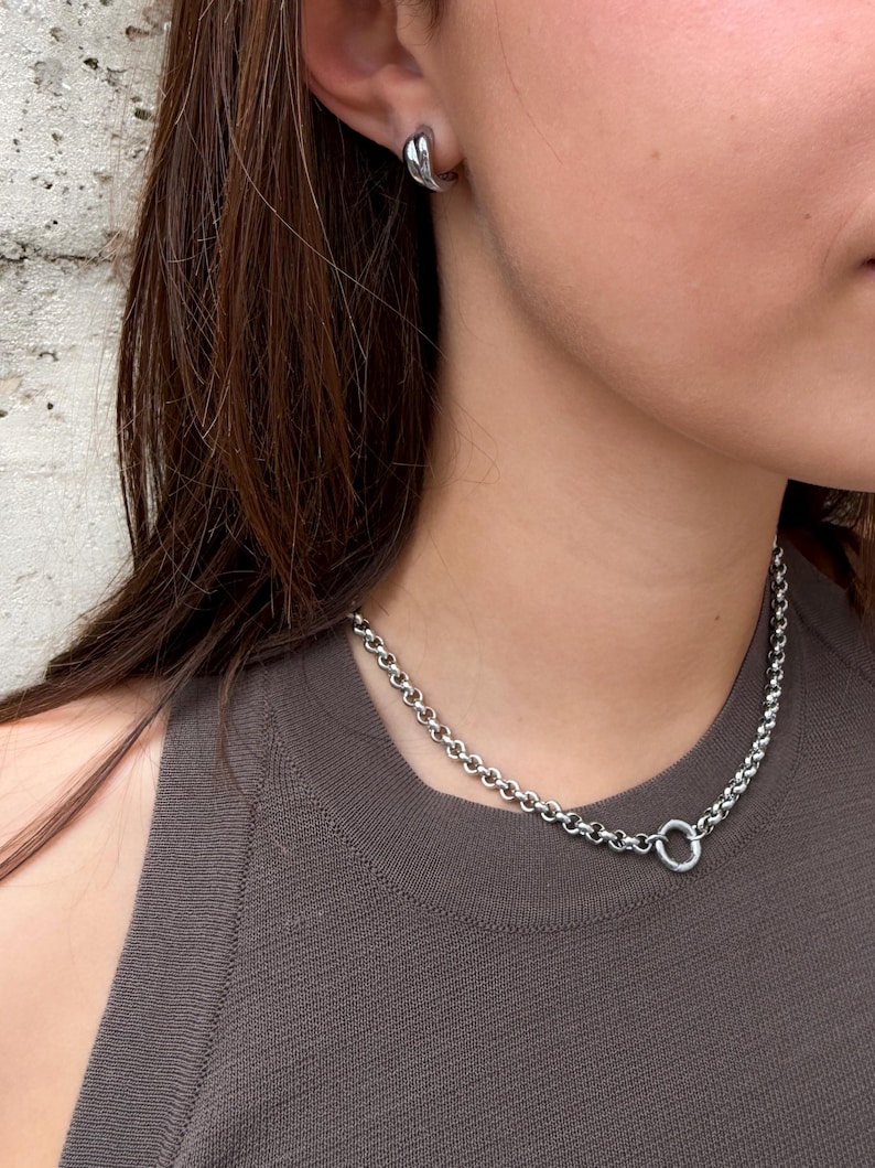 Puede incluir: Un collar de cadena plateada con eslabones redondos y un cierre circular, acompa&ntilde;ado de un pendiente de aro plateado a juego. El collar descansa sobre una camiseta sin mangas de color marr&oacute;n oscuro y el pendiente se lleva en el l&oacute;bulo.