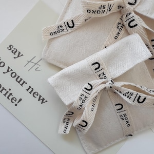 May include: Close-up of three off-white fabric pouches tied with cream-colored ribbons. The ribbons are printed with the word "NONO" and the letter "U" in black. A white card with the text "say Hi to your new favorite!" is in the background.