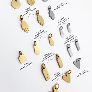 Puede incluir: Una colecci&oacute;n de dijes de metal en tonos oro y plata, con diversas formas que incluyen monedas, manos de hamsa, diamantes, rect&aacute;ngulos, cuadrados y corazones. Algunos dijes son brillantes, otros mate, con algunas piezas grabadas con letras.