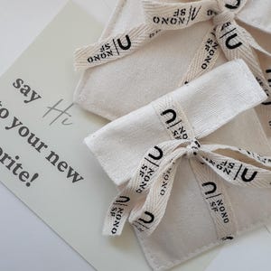 Puede incluir: Dos bolsas de lona color hueso atadas con una cinta color crema que presenta el texto negro "NONO SF" y un logotipo "U". Se ve una tarjeta rectangular con el texto impreso "say Hi to your new favorite!" en una fuente gris oscuro.