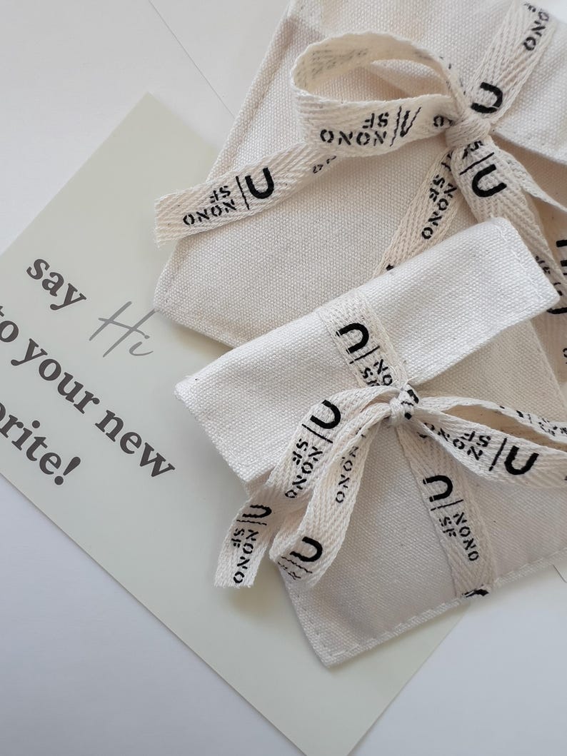 Puede incluir: Dos bolsas de lona color hueso atadas con una cinta color crema que presenta el texto negro "NONO SF" y un logotipo "U". Se ve una tarjeta rectangular con el texto impreso "say Hi to your new favorite!" en una fuente gris oscuro.