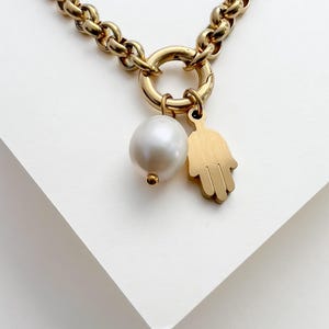 Puede incluir: Un collar de cadena en tono dorado con un cierre circular. Del cierre cuelgan dos dijes: una perla blanca redonda con una peque&ntilde;a cuenta dorada en la base y un dije plano de mano de Hamsa en tono dorado con un acabado liso y mate.