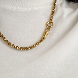 May include: A gold-toned rolo chain necklace featuring a prominent circular link and a swivel lobster clasp closure. The chain is composed of uniform, rounded links that create a continuous, textured appearance against a plain white fabric background.