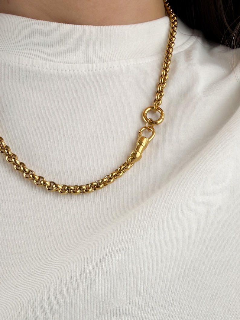 May include: A gold-toned rolo chain necklace featuring a prominent circular link and a swivel lobster clasp closure. The chain is composed of uniform, rounded links that create a continuous, textured appearance against a plain white fabric background.