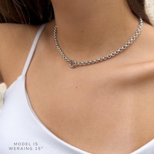 Puede incluir: Un collar de cadena tipo rolo en tono plateado con un cierre de anillo circular posicionado en la parte frontal. El collar mide 38,1 cm de largo, tal como indica el texto en la imagen. La cadena consta de eslabones redondos uniformes.