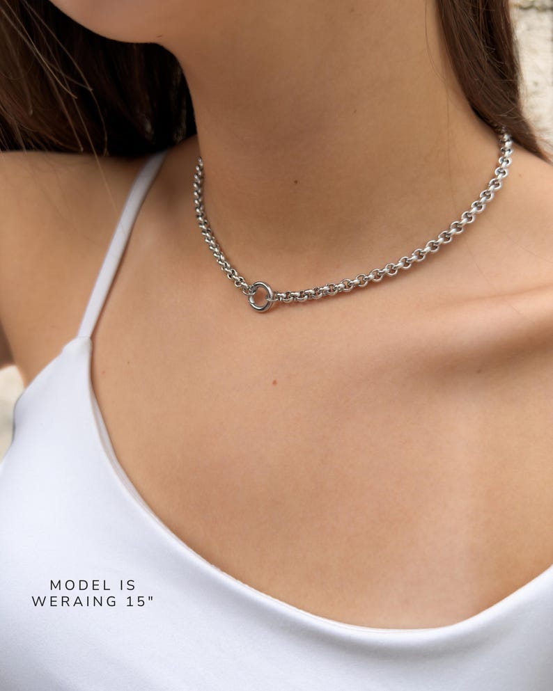 Puede incluir: Un collar de cadena tipo rolo en tono plateado con un cierre de anillo circular posicionado en la parte frontal. El collar mide 38,1 cm de largo, tal como indica el texto en la imagen. La cadena consta de eslabones redondos uniformes.