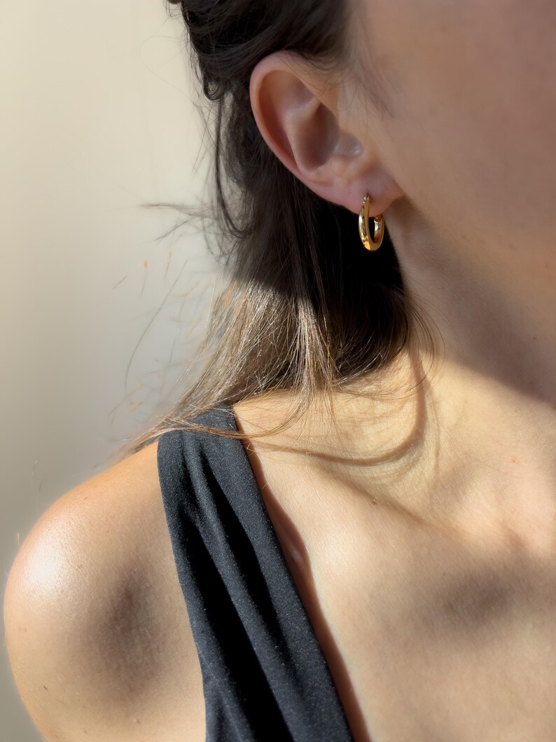 May include: A close-up shot of a gold hoop earring worn on an ear. The earring is a small, circular design with a polished finish. The person is wearing a black tank top.