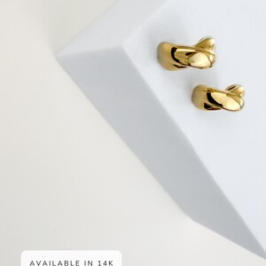 May include: Two gold-plated ear cuffs with a twisted design. The ear cuffs are displayed on a white surface. The text "AVAILABLE IN 14K GOLD PLATED" is visible in a white box.