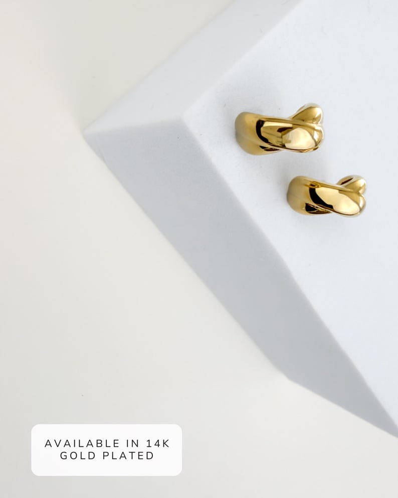 May include: Two gold-plated ear cuffs with a twisted design. The ear cuffs are displayed on a white surface. The text "AVAILABLE IN 14K GOLD PLATED" is visible in a white box.