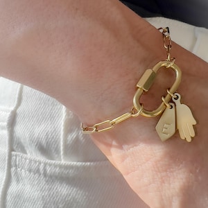 May include: A gold-toned chain bracelet featuring a large oval carabiner-style clasp. Attached to the clasp are two charms: a small, flat, shield-shaped tag engraved with the letter E and a stylised hand-shaped charm with five distinct fingers.