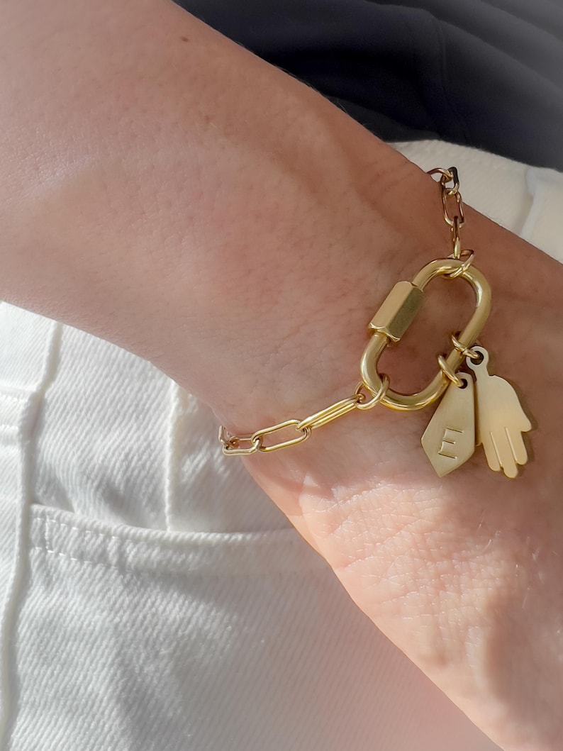 May include: A gold-toned chain bracelet featuring a large oval carabiner-style clasp. Attached to the clasp are two charms: a small, flat, shield-shaped tag engraved with the letter E and a stylised hand-shaped charm with five distinct fingers.