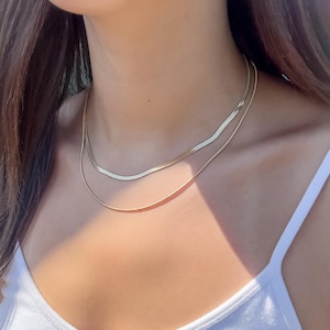 May include: Two gold necklaces of different styles are worn. The top necklace has a flat, herringbone design, while the bottom necklace is a simple, thin chain. The necklaces are worn over a white tank top.