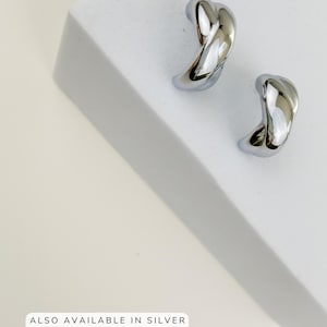 May include: A pair of shiny, silver-colored earrings with a twisted, curved design. The earrings are displayed on a white surface. Text at the bottom reads: ALSO AVAILABLE IN SILVER RHODIUM-PLATED STAINLESS STEEL.