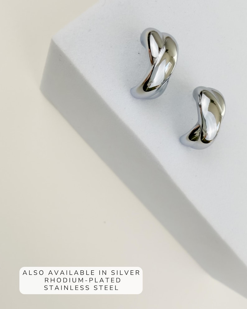 May include: A pair of shiny, silver-colored earrings with a twisted, curved design. The earrings are displayed on a white surface. Text at the bottom reads: ALSO AVAILABLE IN SILVER RHODIUM-PLATED STAINLESS STEEL.