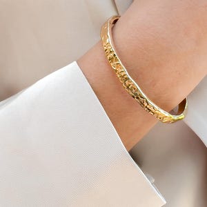 May include: A gold-colored bangle bracelet with a textured surface is worn on a wrist. The bracelet has an organic, hammered design. The person is wearing a light-colored long-sleeved shirt.