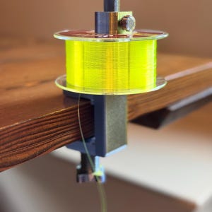 Op de afbeelding: Een spoelhouder voor visdraad met felgele visdraad. De houder is vastgeklemd op een houten oppervlak. De houder heeft een zwart en zilver ontwerp met een transparante plastic spoel.