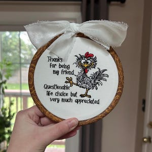 May include: A circular wooden embroidery hoop containing white fabric with a machine-embroidered cartoon chicken and the black text: Thanks for being my friend. Questionable life choice but very much appreciated. A white frayed ribbon is tied at the top.