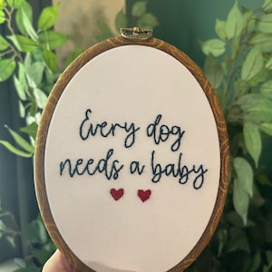 May include: An oval embroidery hoop with a white fabric center. The text "Every dog needs a baby" is stitched in dark blue thread, with two red heart accents. The wooden hoop has a brass clasp.