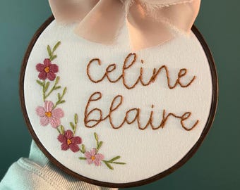 Puede incluir: Arte de aro bordado con los nombres "Celine Blaire" en hilo marrón sobre tela blanca. El diseño incluye flores rosas y burdeos con hojas verdes. Un lazo de cinta rosa claro adorna la parte superior. El aro está enmarcado en madera marrón oscuro.