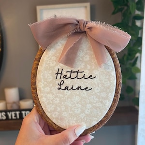 May include: An oval embroidery hoop with a dark wood grain frame containing cream-colored fabric with a subtle white floral pattern. The name Hattie Laine is embroidered in black thread in the center, topped with a dusty rose frayed ribbon bow.