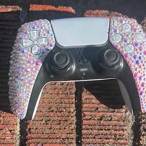 May include: A black and white video game controller with iridescent rhinestones covering the sides. The controller has black joysticks, buttons, and a white center panel. The buttons are clear and the controller has a PlayStation logo.