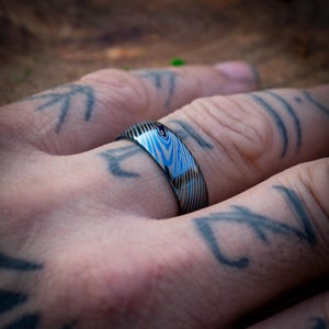 Op de afbeelding: Een blauwe en zilveren ring met een patroon en een geborstelde afwerking. De ring wordt gedragen op een vinger met een tatoeage.
