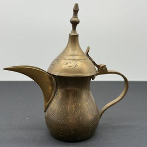 Puede incluir: Una pequeña cafetera antigua de latón con un pico largo y un asa. La cafetera tiene una tapa decorativa y está adornada con patrones florales grabados. El diseño general es ornamentado y tradicional.