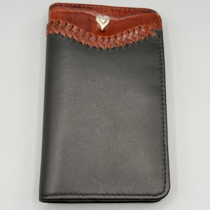 May include: A black leather wallet with a brown, textured top section. The top features a braided design and a silver heart-shaped accent. The wallet is closed and has a simple, elegant design.