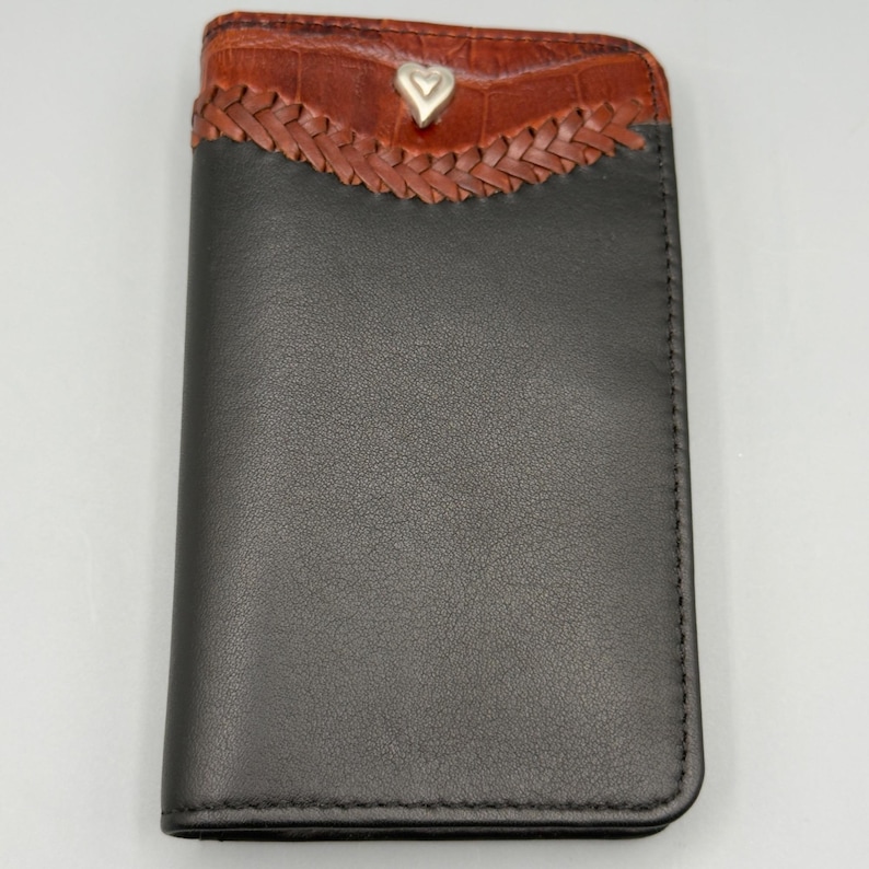 May include: A black leather wallet with a brown, textured top section. The top features a braided design and a silver heart-shaped accent. The wallet is closed and has a simple, elegant design.