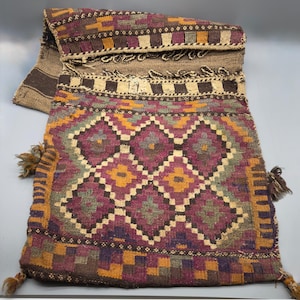 May include: A woven shoulder bag with a geometric pattern in shades of purple, gold, and green. The bag has a flap closure and decorative tassels. The design features diamond and square motifs.