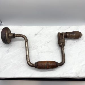 Puede incluir: Un taladro manual antiguo con mango de madera y marco de metal. El taladro tiene un diseño curvo con una cabeza circular en un extremo y un portabrocas en el otro. El mango de madera tiene una etiqueta con texto descolorido.