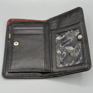 May include: Open black leather wallet with multiple card slots and a clear ID window. The wallet has a snap closure and a textured surface. The interior is black with a brown accent on the top edge.