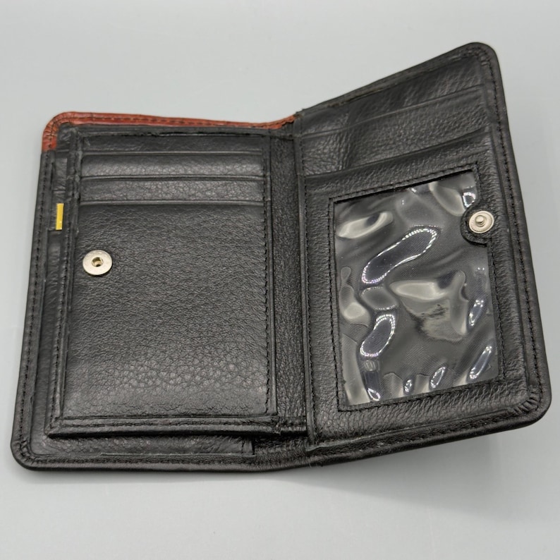 May include: Open black leather wallet with multiple card slots and a clear ID window. The wallet has a snap closure and a textured surface. The interior is black with a brown accent on the top edge.