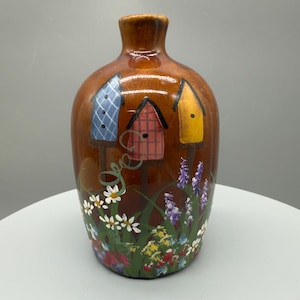 Puede incluir: Un jarrón de cerámica marrón con cuello estrecho, con casas para pájaros pintadas a mano en azul, rojo y amarillo. La base está decorada con flores silvestres blancas, moradas y amarillas, sobre un fondo verde.