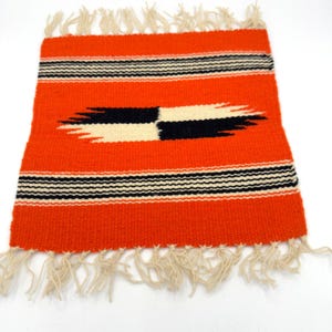 May include: A small, rectangular woven rug with an orange background. The rug features a central design of black and cream-colored shapes, and horizontal black and cream stripes. The edges are finished with cream-colored fringe.