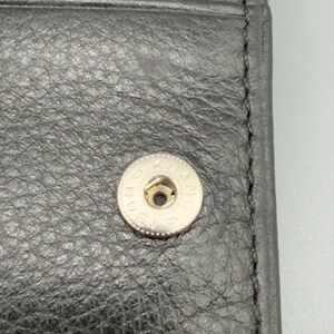 May include: Close-up of a black leather wallet with a silver snap closure. The snap is circular with the text "KUANG" around the edge. The leather has a textured appearance.