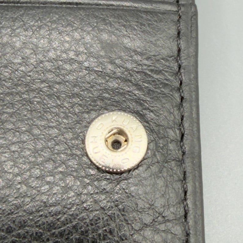 May include: Close-up of a black leather wallet with a silver snap closure. The snap is circular with the text "KUANG" around the edge. The leather has a textured appearance.