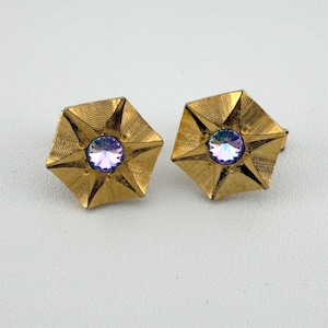 May include: A pair of gold-toned metal cufflinks featuring a hexagonal star-shaped design with a textured surface and a central round, faceted, iridescent light purple stone set in the middle of each piece, creating a reflective appearance.