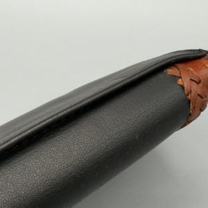 May include: Close-up of a black leather object with a textured surface. The edge is accented with brown leather and a woven design. The object appears to be a wallet or a similar accessory.