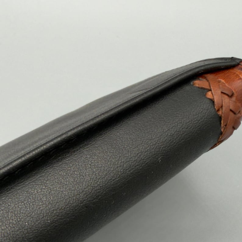 May include: Close-up of a black leather object with a textured surface. The edge is accented with brown leather and a woven design. The object appears to be a wallet or a similar accessory.