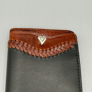 May include: A black leather wallet with a brown, textured top section. The top features a woven design and a silver heart-shaped emblem. The wallet is a rectangular shape.