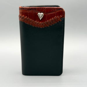 May include: Black leather wallet with a decorative top section. The top features a brown, textured material with a woven design and a silver heart-shaped accent. The wallet is rectangular and appears to be new.