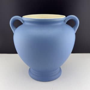 May include: A blue ceramic vase with two curved handles. The vase has a wide body and a narrow base. The interior rim is a cream color. The vase is a matte finish and sits on a white surface.