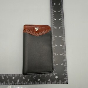 May include: Black leather card holder with a brown, braided top section. The top section has a brown, textured leather trim and a small, silver heart-shaped emblem. The card holder measures approximately 4 inches in height.