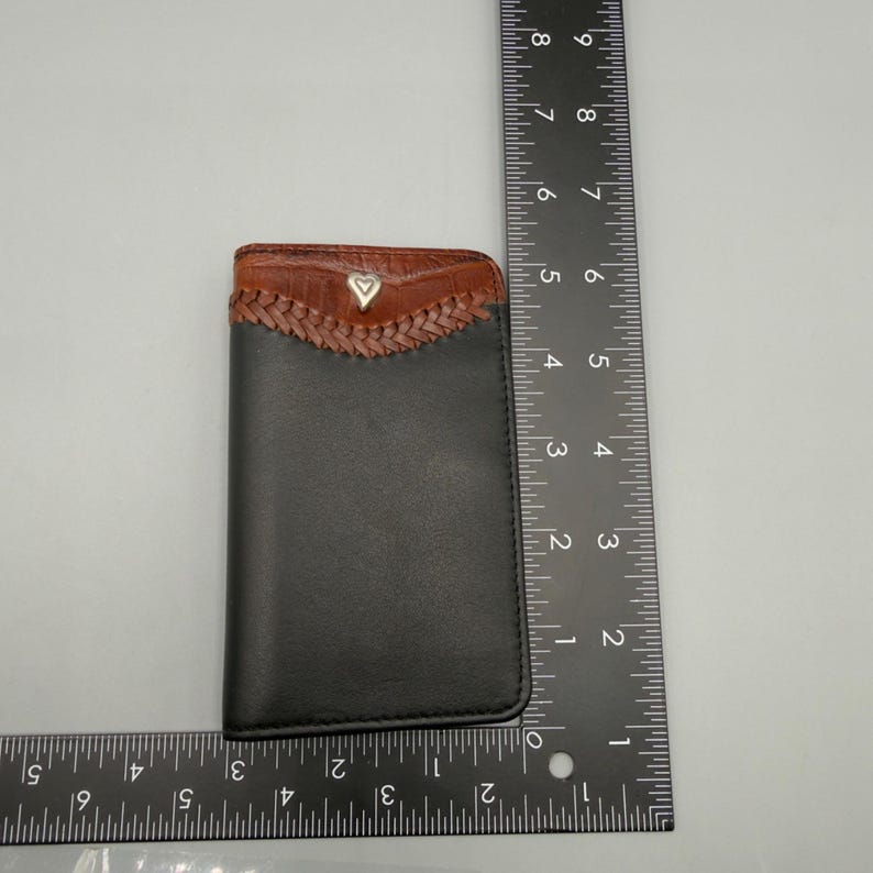 May include: Black leather card holder with a brown, braided top section. The top section has a brown, textured leather trim and a small, silver heart-shaped emblem. The card holder measures approximately 4 inches in height.
